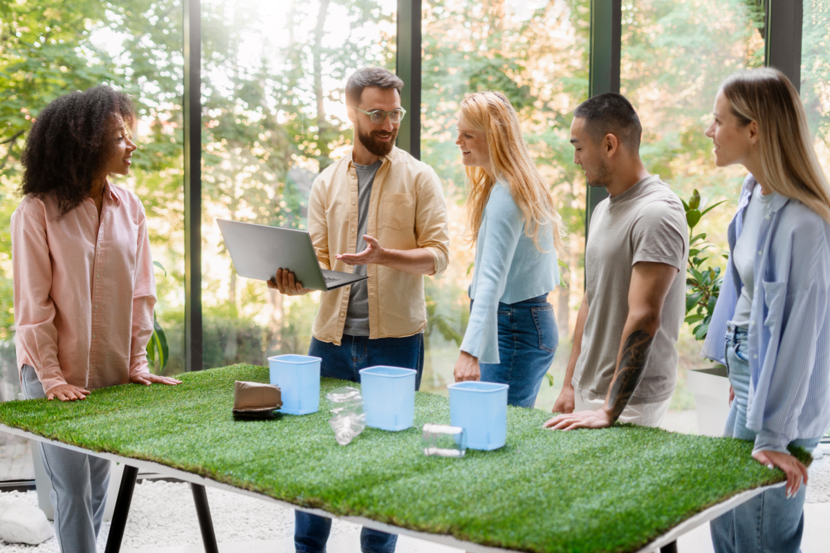 Group of five young adults discussing around a grass-covered table with recycling bins and plastic waste, in a bright, modern room.